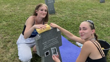 Zwei Teilnehmerinnen hocken neben einem Grabstein. Eine von ihnen hält ein Bild von einem jungen Mann in der Hand, welches am Grabstein befestigt war. Auf dem Grabstein steht der Name Karl Steinhauer zusammen mit dem Verweis auf drei unbekannte deutsche Soldaten.
