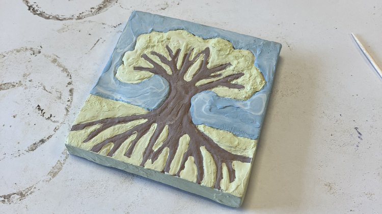 Schräg auf einem weißen Blatt Papier liegt eine bemalte Tonkachel. Sie zeigt einen Baum, dessen Wurzeln tief in den Boden reichen, er ist von einem hellblauen Himmel umgeben.