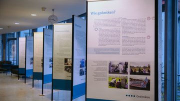 Blick auf vier hintereinander in Reihe aufgebaute Metallgestelle, an welche die Stoffbanner der Ausstellung montiert wurden. Auf dem Element im Vordergrund ist die Fragestellung "Wie gedenken?" zu erkennen. Darunter Text und verschiedene Bilder.