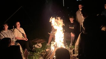 Um eine Feuerquelle (Lagerfeuer) sitzt bei Dunkelheit eine Gruppe junger Menschen versammelt. Die nachdenklichen Gesichter der Teilnehmenden im Hintergrund sind durch den Schein des Feuers erkennbar.