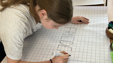 Eine Schülerin steht links im Bild nach rechts über ein kariertes A1-Papier auf einem Tisch gebeugt. Sie schreibt mit einem schwarzen Edding-Stift das Wort "Frieden" in Großbuchstaben.