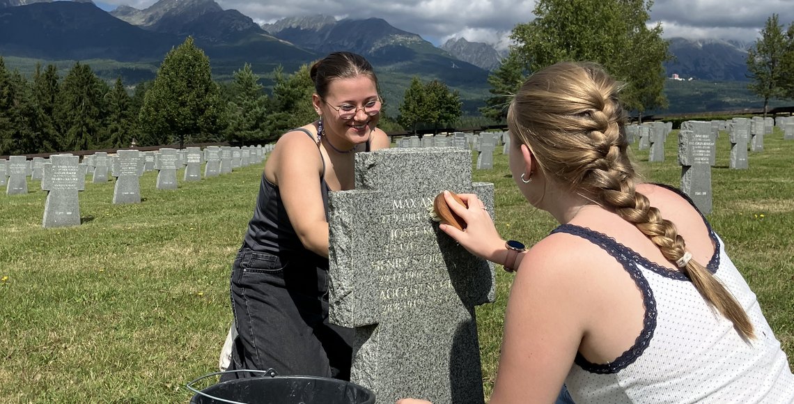 Zwei Teilnehmerinnen der Jugendbegegnung sitzen vor und hinter einem Grabstein auf einem Friedhof. Sie benutzen Bürsten, den Stein von Moosen und Flechten zu reinigen. Im Hintergrund: Wald und eine slowakische Berglandschaft.