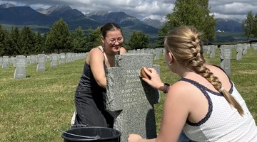 Zwei Teilnehmerinnen der Jugendbegegnung sitzen vor und hinter einem Grabstein auf einem Friedhof. Sie benutzen Bürsten, den Stein von Moosen und Flechten zu reinigen. Im Hintergrund: Wald und eine slowakische Berglandschaft.