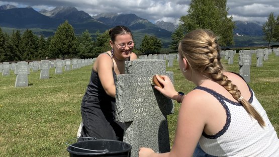 Zwei Teilnehmerinnen der Jugendbegegnung sitzen vor und hinter einem Grabstein auf einem Friedhof. Sie benutzen Bürsten, den Stein von Moosen und Flechten zu reinigen. Im Hintergrund: Wald und eine slowakische Berglandschaft.