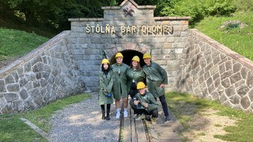 Fünf Teilnehmende stehen vor einem Stolleneingang mit dem goldenen Schriftzug "Štôlňa Bartolomej". Er ist Teil des Bergbaumuseums in Banská Štiavnica. Alle fünf Personen tragen grüne Regenmäntel und gelbe Helme.