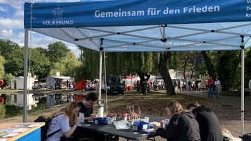 Je zwei junge Menschen sitzen zu beiden Seiten eines Tisches einander gegenüber überdacht von einem hellblauen Pavillon mit der Aufschrift "Volksbund - Gemeinsam für den Frieden". Am Tisch gestalten und bemalen sie Friedenskacheln. Im Vordergrund auf der linken Seite ein Stand mit Info-Flyern.