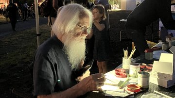 Ein Mann mit weißen Haaren und Vollbart sitzt in der Bildmitte auf einer Bank überdacht von einem Pavillon im Dunkeln. Vor ihm auf der rechten Seite steht ein Tisch, auf dem er im Licht seiner Handy-Taschenlampe eine Friedenskachel bemalt.