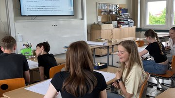 Schülerinnen und Schüler sitzen in einem Klassenzimmer in Paaren über mehrere Sitzreihen verteilt. Vor ihnen liegt Papier im A1-Format, welches sie beschriften. Im Hintergrund befindet sich eine digitale Tafel, welche die Aufgabenstellung anzeigt.