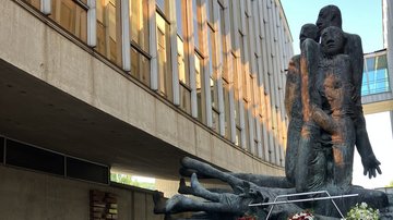 Modernes Kunstwerk im Zentrum eines Innenhofs eines Museums. Vor ihm sind zahlreiche Kränze abgelegt. Oberhalb ist eine Beton- und Fensterfassade zu erkennen, welche zu einer Hälfte des Museumsgebäudes gehört.