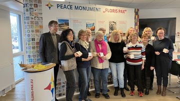 Vor einer Stellwand steht eine Gruppe von 13 Frauen und einem Mann. Sie lächeln für ein Gruppenbild in die Kamera und halten ihre Tonkacheln in Richtung der Kamera. Hinter ihnen befindet sich eine bunt gestaltete Stellwand mit dem Schriftzug "Peace Monument Floris Pax".