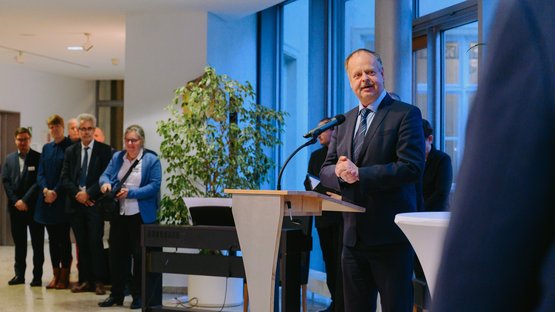 Der Landtagsvizepräsident steht festlich gekleidet am Rednerpult vor einer Fensterfront und blickt in Richtung der Kamera. Im Hintergrund steht eine Gruppe von acht Personen, die zum Publikum gehören.