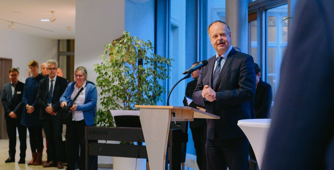 Der Landtagsvizepräsident steht festlich gekleidet am Rednerpult vor einer Fensterfront und blickt in Richtung der Kamera. Im Hintergrund steht eine Gruppe von acht Personen, die zum Publikum gehören.