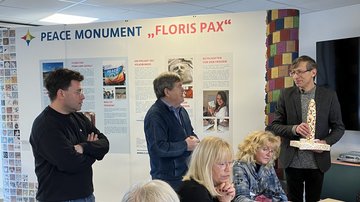 Drei Männer stehen nebeneinander vor einer Stellwand mit der Beschriftung "Peace Monument Floris Pax". Vor ihnen sitzt eine Gruppe Frauen an einem Tisch und hört den Ausführungen zu.