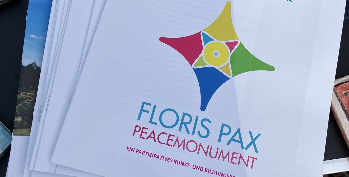 Draufsicht von oben auf verschiedene Flyer und Infomaterialien auf einem schwarzen Tisch. Oberhalb davon kleine quadratische Tonkacheln mit dem Schriftzug "Frieden" sowie einer Katze und einer Sonnenblume.