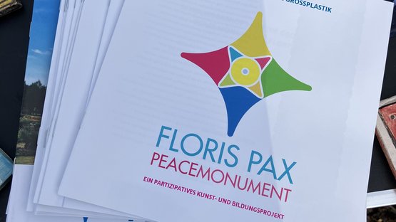Draufsicht von oben auf verschiedene Flyer und Infomaterialien auf einem schwarzen Tisch. Oberhalb davon kleine quadratische Tonkacheln mit dem Schriftzug "Frieden" sowie einer Katze und einer Sonnenblume.